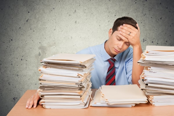 man with head in hand behind stacks of folders and papers
