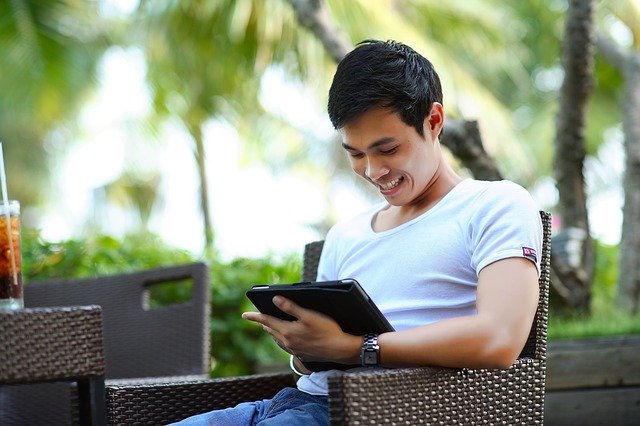 man in t-shirt sits at table looking at tablet and smiles