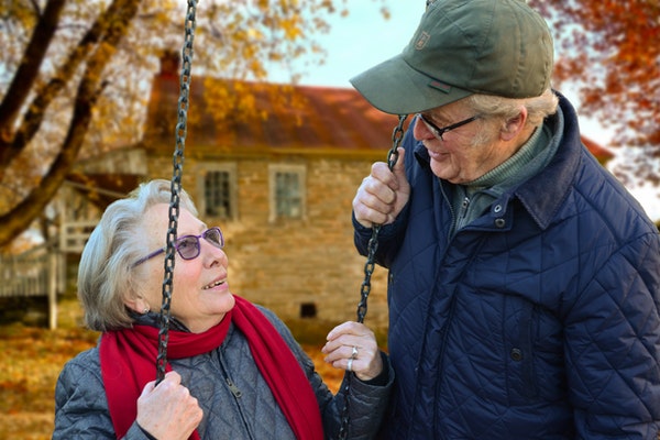 old couple smile at each other on a swing in front of house