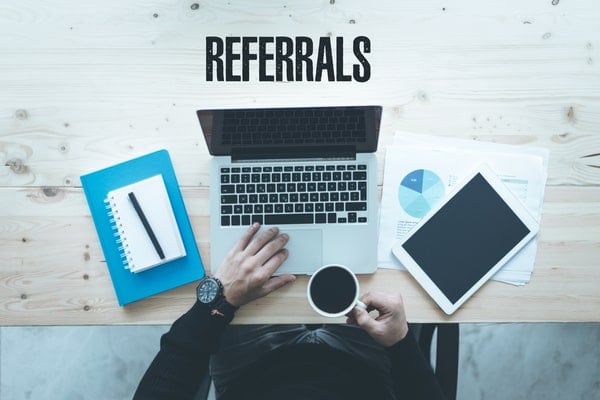 man at a table with the word "referrals", laptop, coffee mug, and notebook